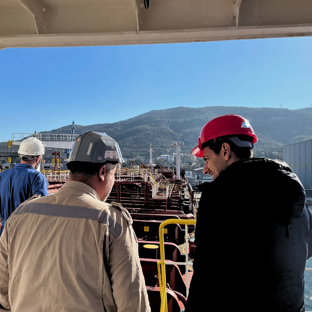 Katradis Maritime engineers conducting an on-site inspection of port infrastructure and vessel safety systems, wearing protective gear and hard hats, with a backdrop of a modern port and surrounding hills.