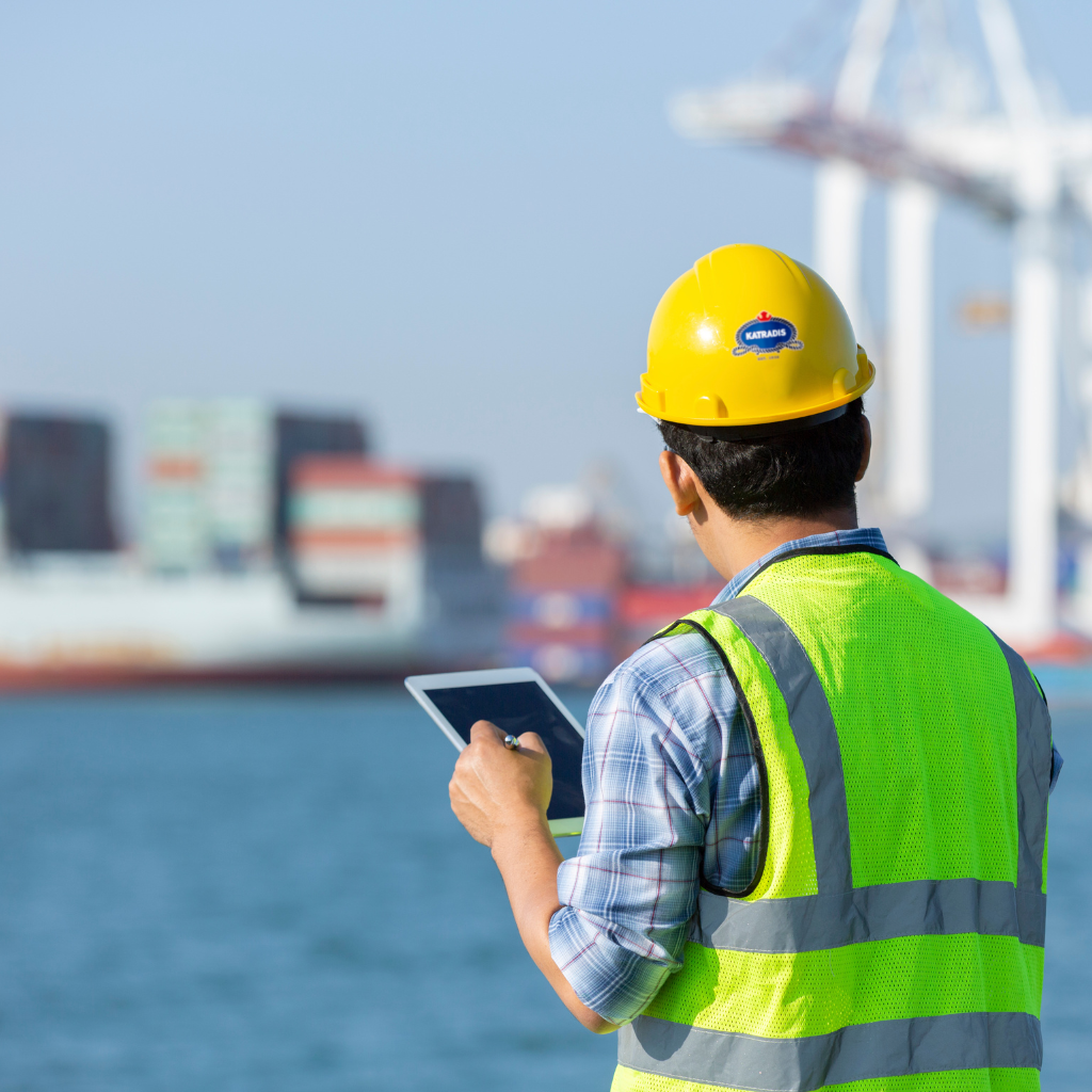 Marine engineer conducting a port infrastructure assessment using a tablet, wearing safety gear including a hard hat and high-visibility vest, with container ships and cranes in the background.